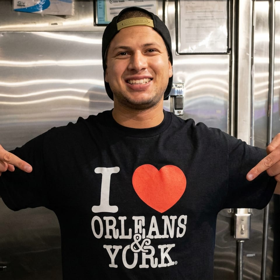 Smiling restaurant worker points to an “I love Orleans & York” shirt inside a stainless-steel kitchen.