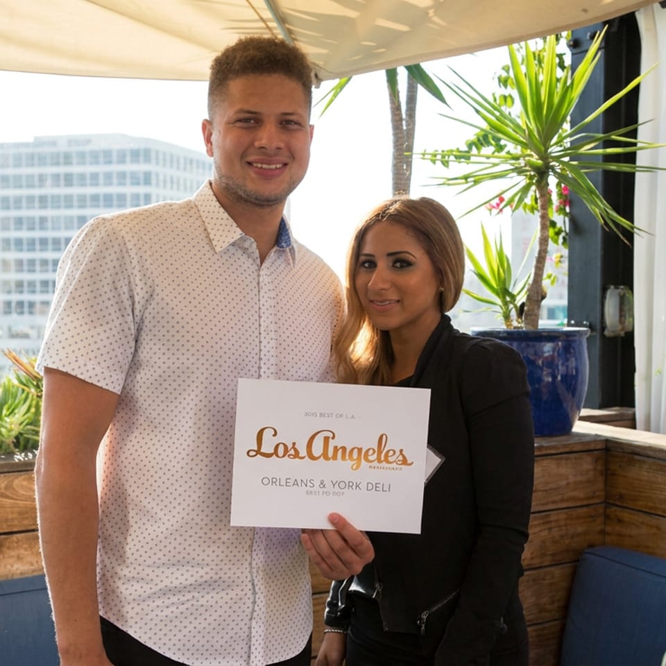 Two people pose on a sunny rooftop patio, holding a Los Angeles award sign for Orleans & York Deli.