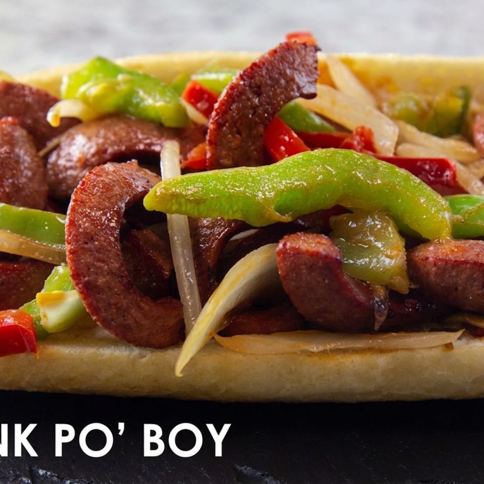Hot link po’ boy sandwich on a French roll, topped with sliced sausage, green peppers, red peppers, and onions.