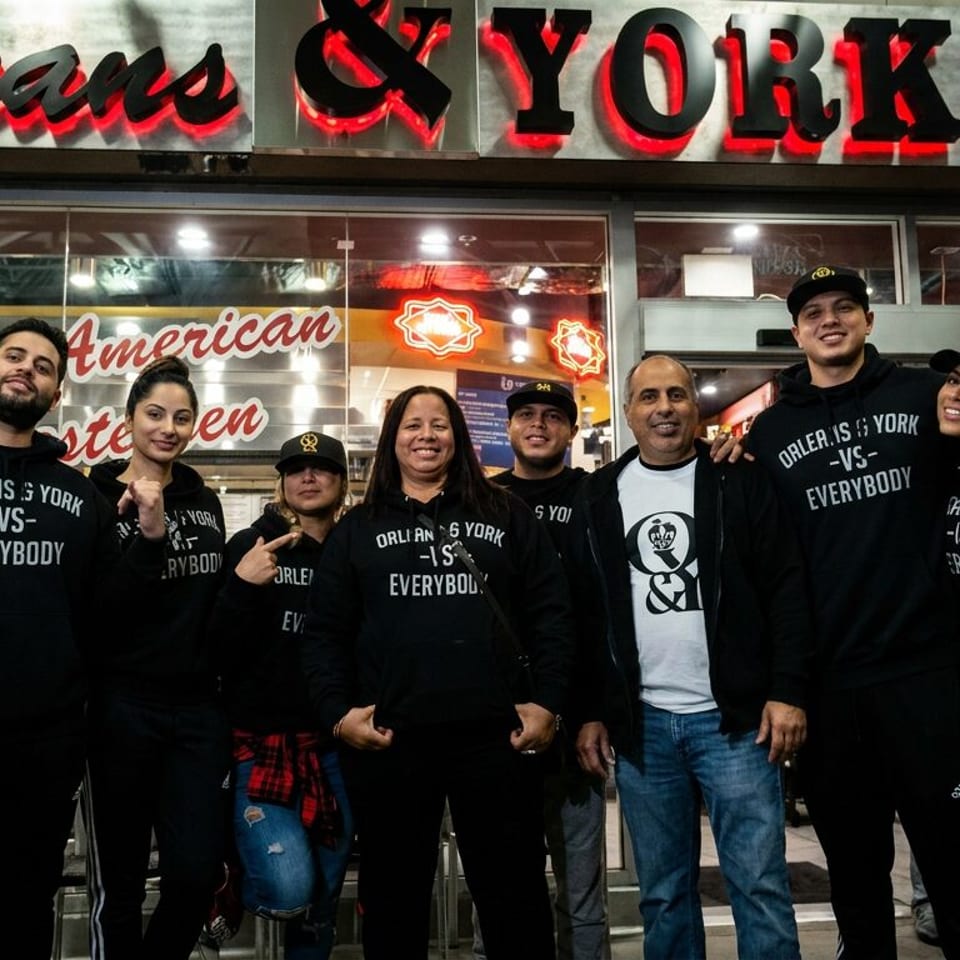 Group of seven people wearing Orleans & York apparel pose outside the restaurant beneath its illuminated sign.