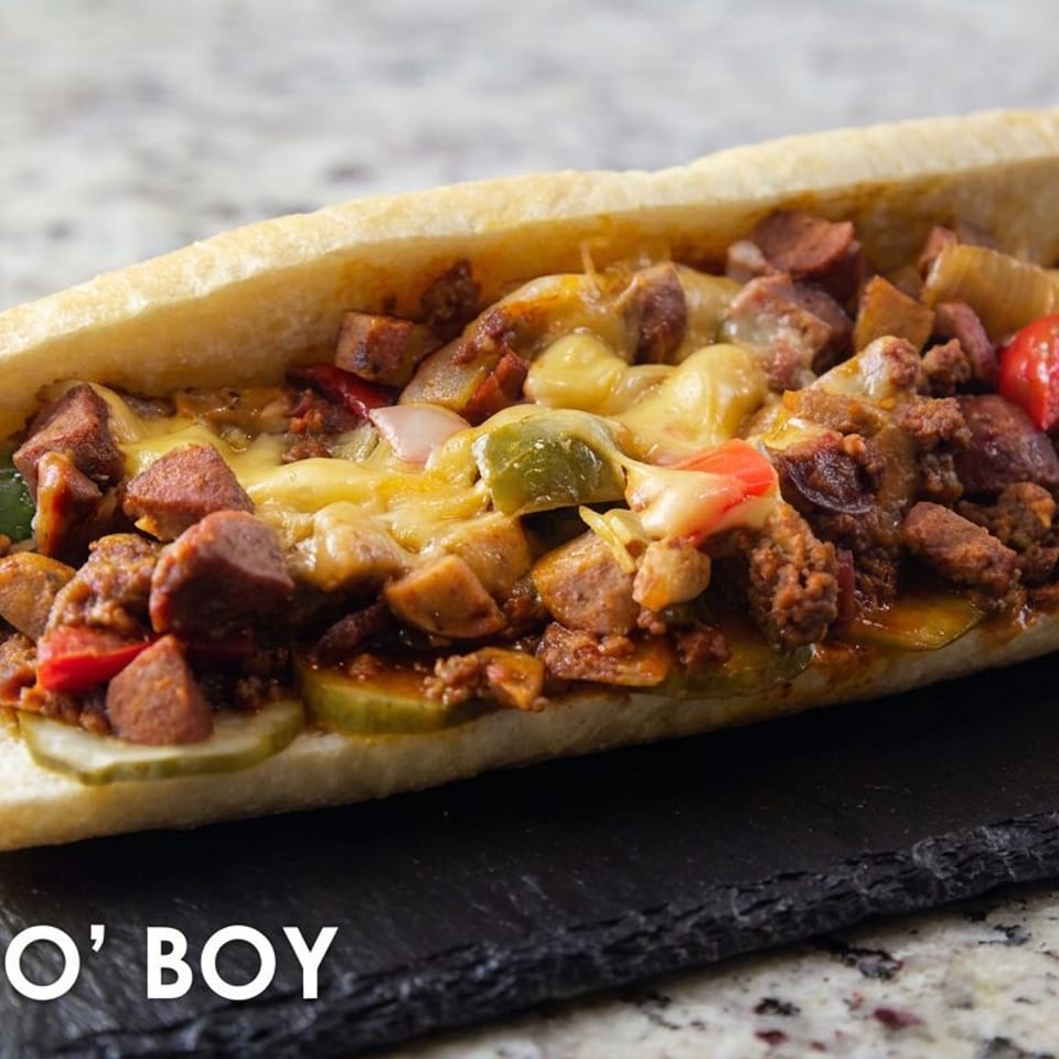 Loaded Dirty Po’ Boy sandwich with seasoned meat, sausage, peppers, pickles, and melted cheese on a slate serving board.