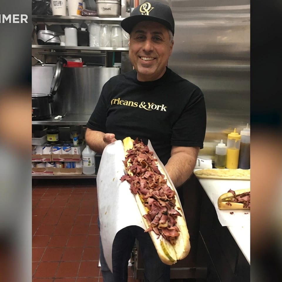 Restaurant worker presents an oversized hot dog piled with sliced meat in a commercial kitchen.